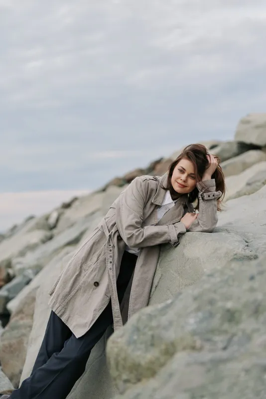 Young woman in knitted coat leans on large rocks outdoors under cloudy sky. Casual fashion concept with natural makeup and relaxed mood in a coastal environment.