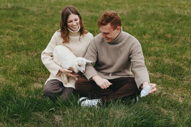 Couple sits on a grassy field holding a white goat kid, smiling and relaxed, enjoying a calm outdoor moment in cozy sweaters, warm light, lifestyle portrait showing affection and connection.