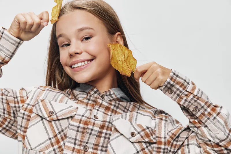Girl smiling with autumn leaves near face wearing plaid shirt, posing with happiness and playful mood in casual cozy outfit for seasonal outdoor portrait.