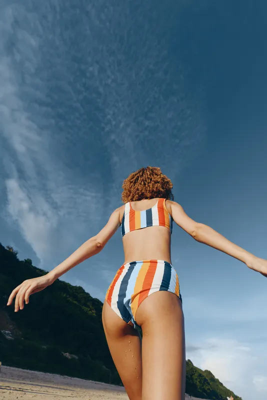 Woman, striped swimsuit, back view, arms outstretched, beach, sky, summer, freedom, vacation, seaside outdoors with bright sunlight, clouds, coastal landscape, relaxed confident posture