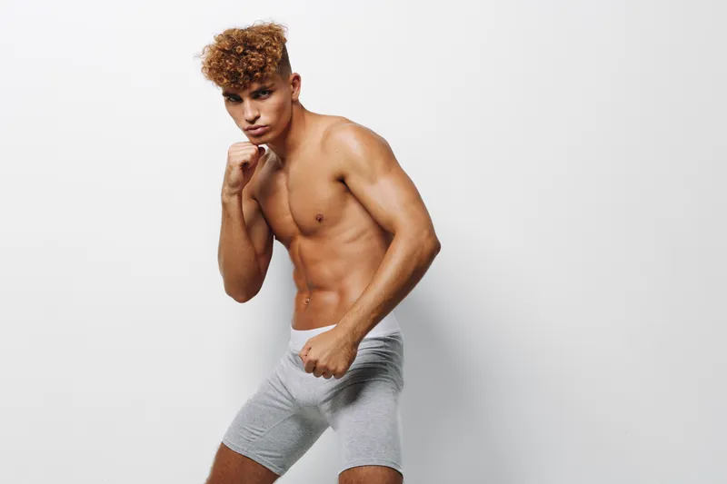 Young man with curly hair posing barechested against white background wearing gray shorts with tors and arms tense muscular fit model with confident facial expression.