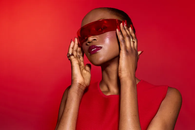 Stylish African woman with dark skin and bald head poses confidently wearing red futuristic glasses and sleeveless dress against a vibrant red background. Fashion and beauty concept.