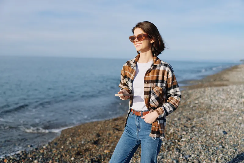Casual woman by the sea on a sunny day, wearing a plaid shirt and jeans, holding a smartphone, enjoying a relaxed coastal stroll with sunglasses and ocean breeze.