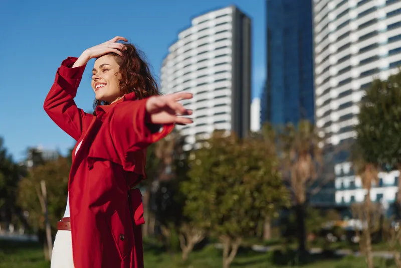 Cheerful woman outdoors wearing a red coat, smiling with bright eyes, arms raised, breeze in hair, urban park setting with tall buildings and clear sky blue.