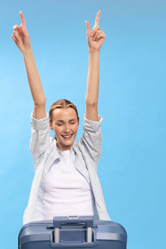 Excited young woman with raised arms and closed eyes stands behind blue suitcase on blue background. Concept of travel, vacation mood, happiness, and freedom before journey.