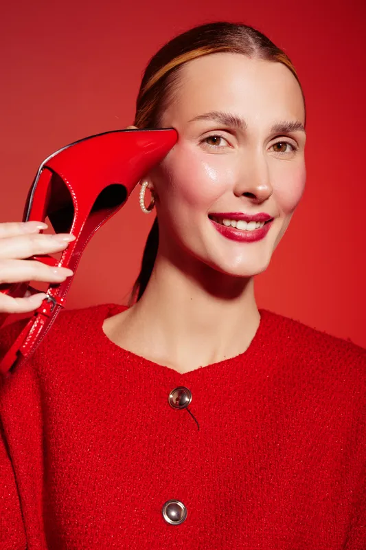 Smiling woman in red sweater holds red high heel shoe near face on red background. Fashion concept with bright colors, beauty, and stylish accessories for modern lifestyle and elegance.