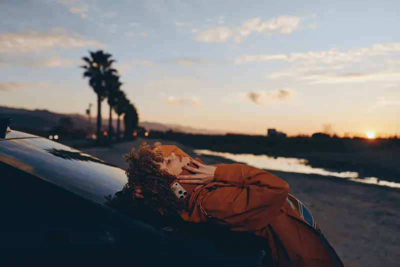 Woman lifestyle portrait at sunset on a beach, she leans on a car hood with relaxed pose, ocean waves and palm trees in soft evening light, travel mood, calm lifestyle concept