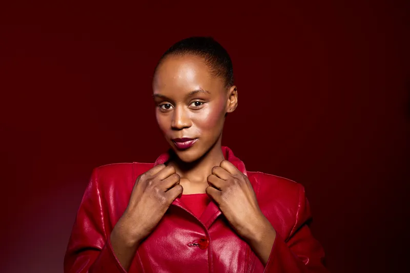 Confident black woman with dark skin wearing red leather jacket poses against dark red background. Studio portrait of African American female model showcasing fashion and style concept.