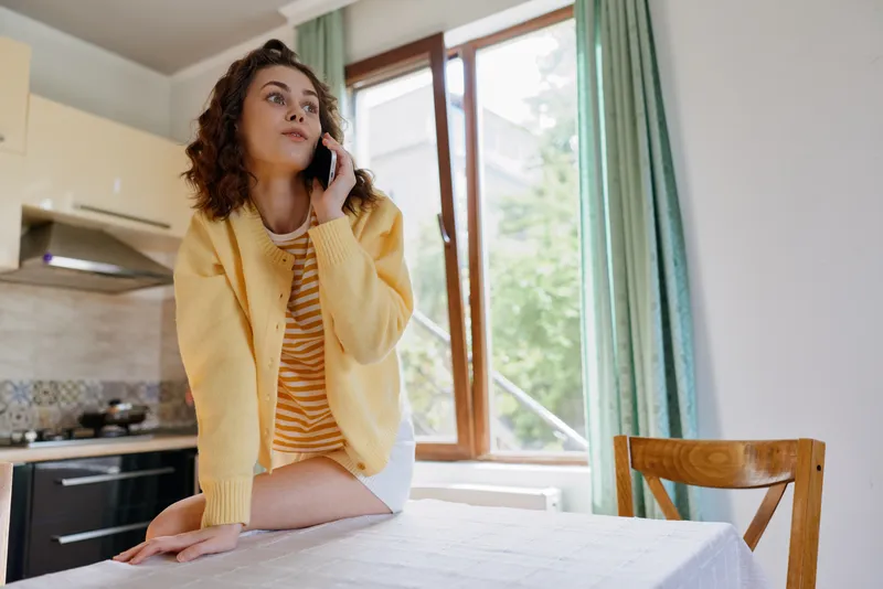 Cheerful woman indoors, talking on phone in bright kitchen, warm yellow cardigan, striped dress, relaxed posture, home lifestyle vibe, natural light, cozy atmosphere, contemporary domestic scene