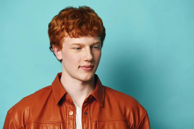 Young man with red hair wearing brown leather jacket standing against blue background looking downwards with calm expression and casual style clothing.