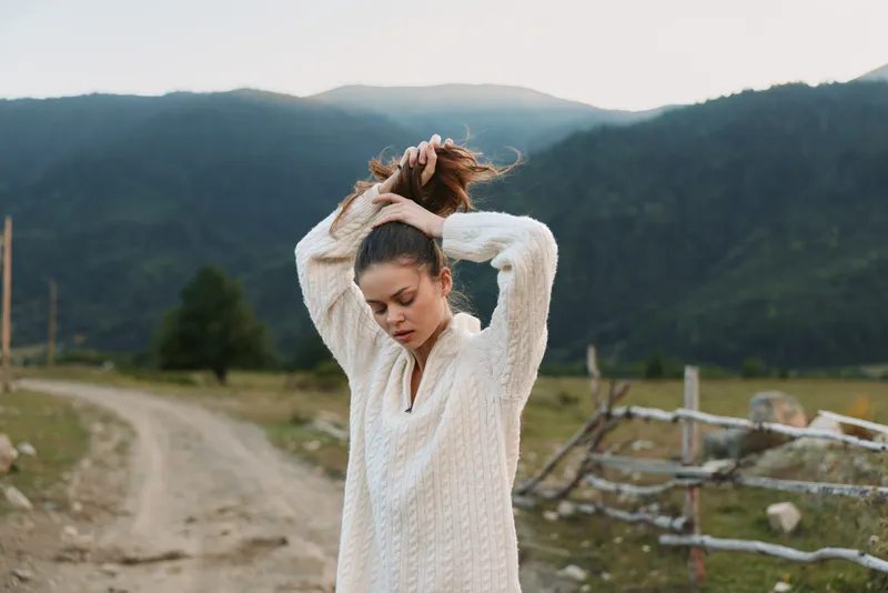 A young woman in a white knit sweater stands on a dirt road in a mountain valley, tying her hair up as natural light highlights the rural landscape.