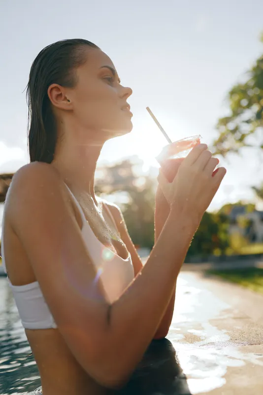 Vibrant lifestyle moment featuring a woman by a sunlit pool, sipping a drink through a straw, relaxed posture, warm glow, reflective water, cheerful mood, casual outdoor summer scene