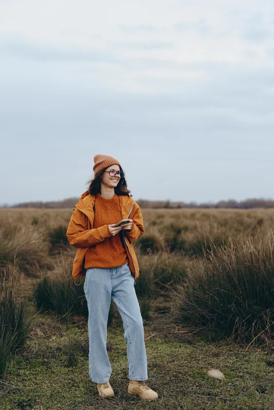 Young woman lifestyle outdoors in countryside field, wearing hat and jacket with jeans, walking while holding phone, smiling. Grassland and cloudy sky behind, orange sweater, scarf, light, pose.