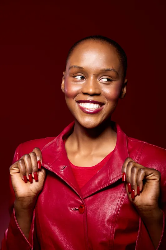 Smiling African American woman with dark skin in red leather jacket poses confidently against red background. Concept of fashion, beauty, confidence, and stylish urban portrait.