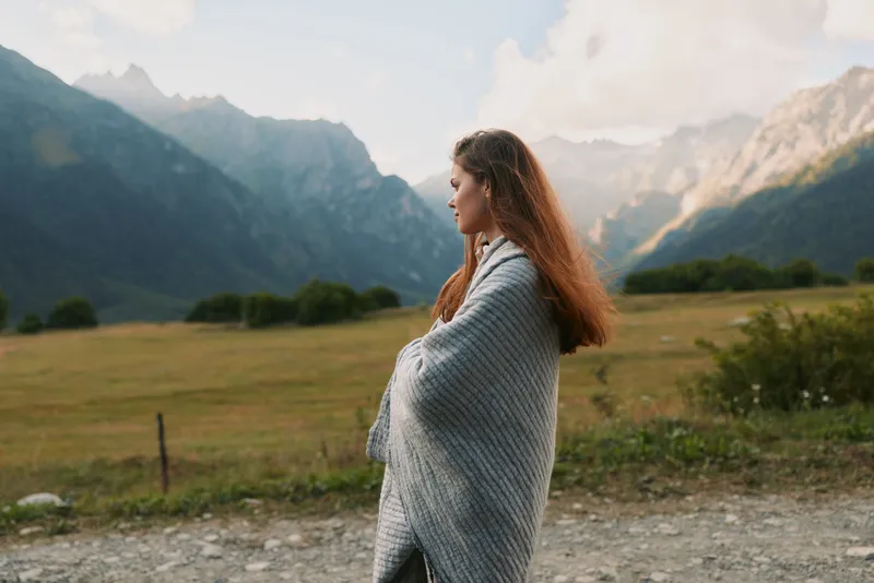 A woman wrapped in a gray blanket stands outdoors in a mountainous landscape, looking contemplative. The scene captures lifestyle and nature connection.