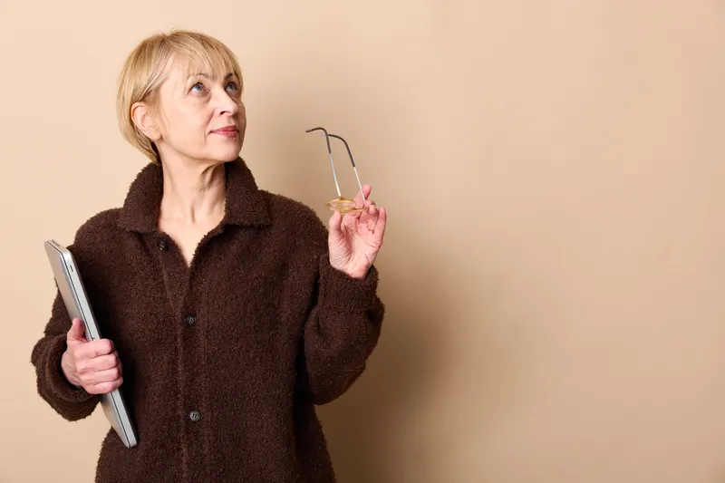 elderly woman studio portrait indoors, beige background, holding eyeglasses and notebook, thoughtful expression, mature age, soft studio light, neutral tone, calm stance, copy space