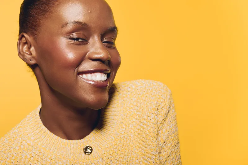 Smiling African American woman with dark skin wearing yellow sweater poses against yellow background. Studio portrait captures positive mood, beauty, fashion, and confidence of young black female.