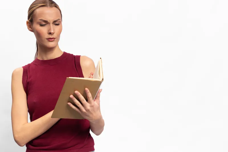 Focused young woman in red sleeveless top reads a book intensely against white background. Concept of concentration, education, learning, and self-development in a minimalistic setting.