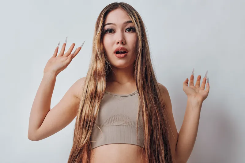 Woman with long hair and casual top standing with raised hands, expressing surprise or shock, isolated on white background in studio portrait.