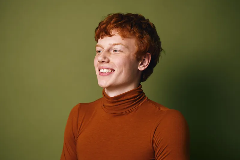 Young man with red hair smiling and looking away wearing orange turtleneck sweater on green background in casual fashion portrait.