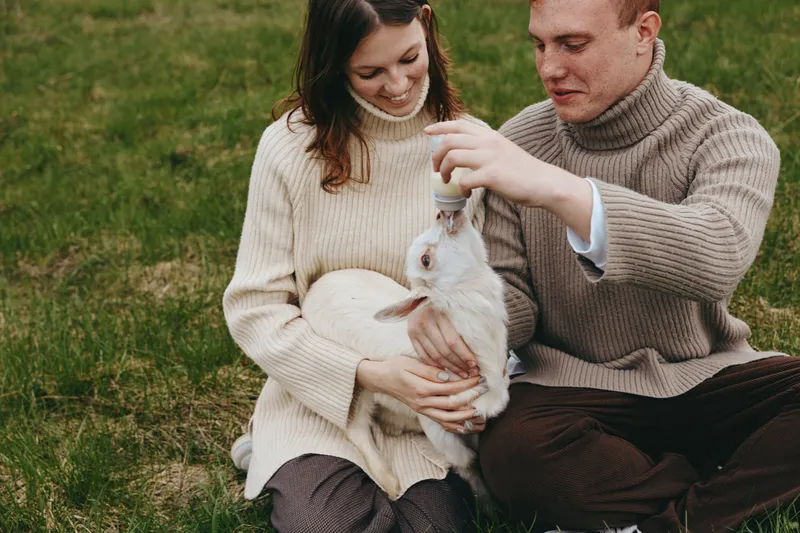 Young couple in warm sweaters feed a small white baby goat while sitting on grass during a cozy outdoor lifestyle moment in autumn.