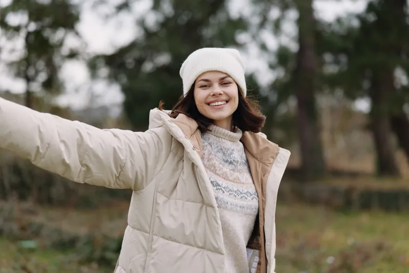 Winter scene with a smiling woman in a padded coat and beanie, arms outstretched in a park, conveying warmth, freedom and casual outdoor style.