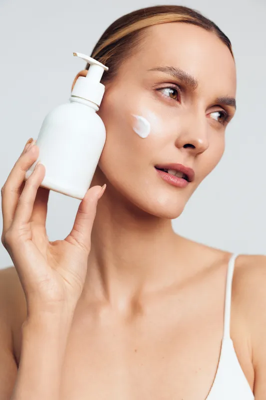 A calm woman applies moisturizer from a pump bottle, highlighting a skincare routine and self care in a bright studio. Clean background, soft lighting, and healthy skin focus, conveying routine care