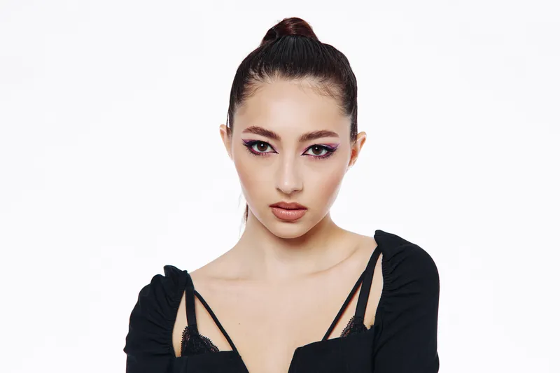 Young woman with creative colorful eyeshadow poses confidently in a black fashion outfit on white background. Concept for beauty, fashion, makeup, and creative styling.