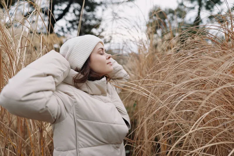 Winter woman in a warm coat and beanie stands among tall grasses, calm and contemplative in a cold outdoor landscape.
