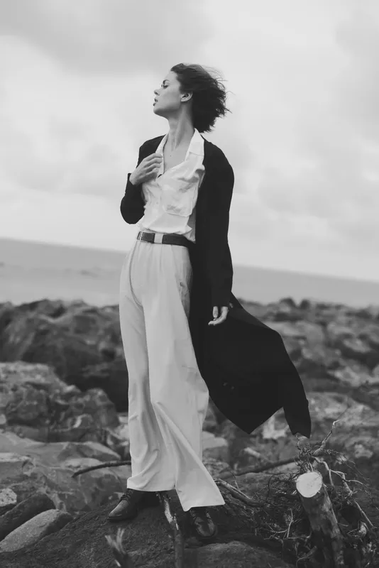 Windy coastal fashion scene, a woman in flowing coat and wide trousers gazing toward the sea on rugged rocks in monochrome.