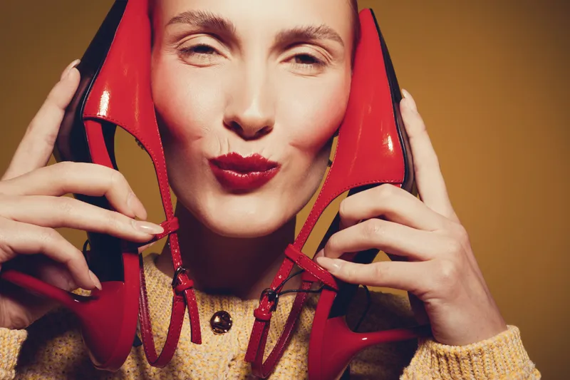 Smiling woman holds red high heel shoes near face, puckering lips. Close-up portrait with warm lighting on brown background. Concept of fashion, style, and beauty accessories.