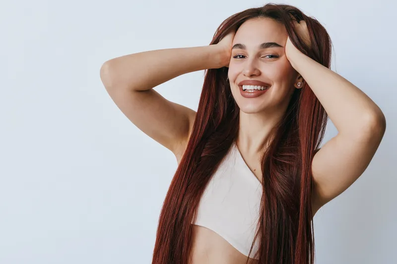 Young woman with long braided hair smiling and posing with hands on head in casual sleeveless top against white background. Beauty, happiness, and confidence in studio portrait.