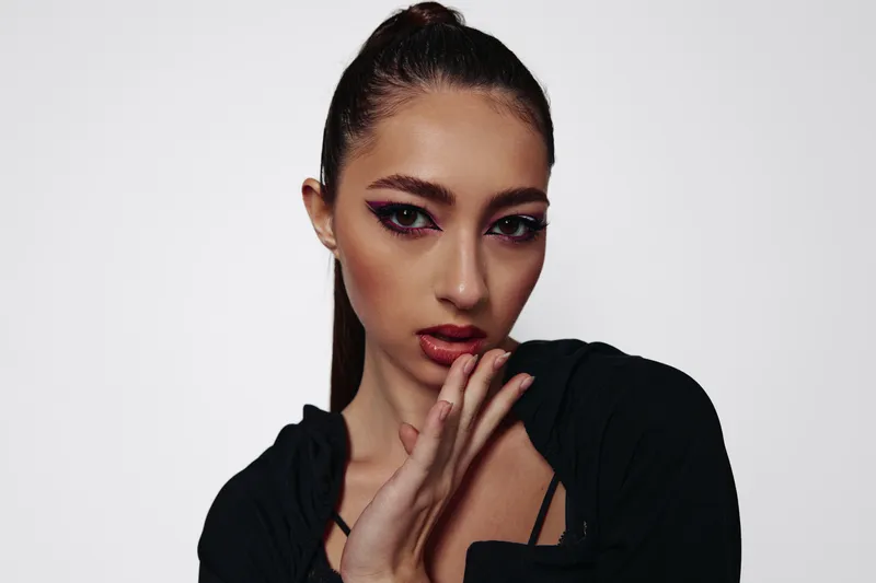 Young woman with creative makeup looks confidently at the camera while gently touching her face. Minimalist background enhances focus on her fashion style and beauty concept.