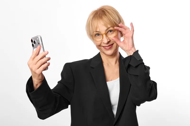 Older woman in business blazer holds smartphone for selfie, wears glasses, waves toward camera, smiling, studio portrait on white background, communication moment