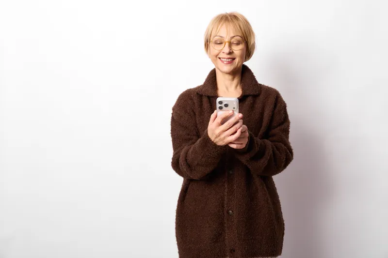 Senior woman portrait, smiling, holding smartphone in her hands, brown winter coat, warm eyeglasses, studio light on white background, casual indoor lifestyle, calm daily routine