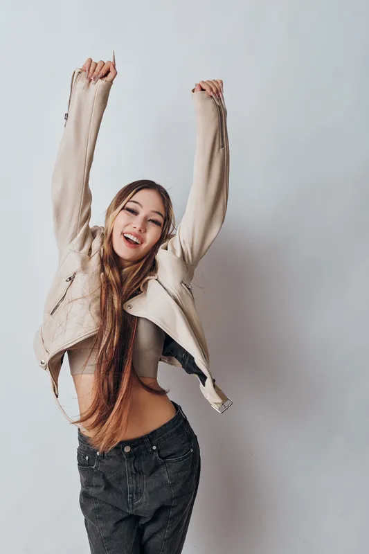 Happy woman with long hair wearing jacket and crop top smiling and stretching arms up against plain background, casual style and positive emotion for lifestyle concept.