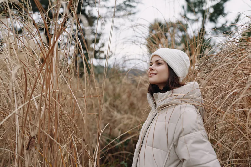 Woman in a beige coat and knit beanie stands among tall dried grass, enjoying a calm outdoor moment. Portrait in natural light with autumn winter scenery and relaxed fashion vibe.