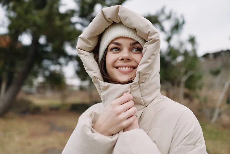 Woman in a warm hooded jacket smiling outdoors in a chilly setting, cozy winter scene with trees and a soft natural background.
