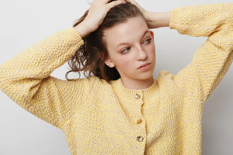 Young woman in yellow knitted sweater adjusts her hair with both hands, looking to the side. Studio portrait on light background. Concept of casual style and natural beauty.