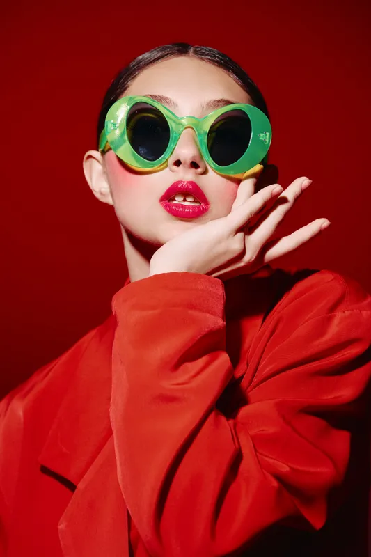 Stylish woman in oversized green sunglasses and bright red coat looks confident posing with hand near face. Portrait on red background. Concept for beauty, fashion, creativity, glamour.