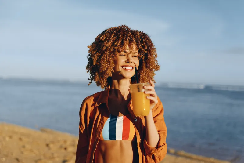 Smiling woman with curly hair on beach by ocean, holding a drink cup and wearing a casual jacket, warm sunlight, relaxed vacation mood, outdoors with soft horizon blur behind.