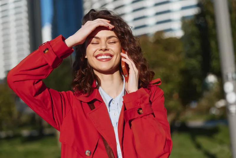Happy smiling woman in a red jacket stands outdoors in a sunny city, talking on a phone, cheerful mood, daylight, urban park setting, candid portrait, joyful moment, bright background