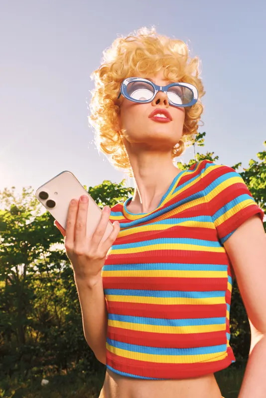 A woman in a striped top and retro sunglasses stands outdoors, capturing a vibrant lifestyle moment with a smartphone, projecting confidence, playfulness, and bold fashion in bright, sunlit film