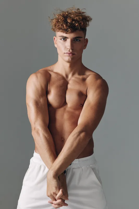 Muscular young man posing shirtless with defined chest and arms wearing white shorts against gray background. Fitness, strength and bodybuilder concept with confident expression and curly hair.