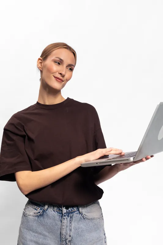 Young woman in casual clothes holds laptop and looks sideways with slight smile on white background. Concept for remote work, technology, and modern lifestyle.