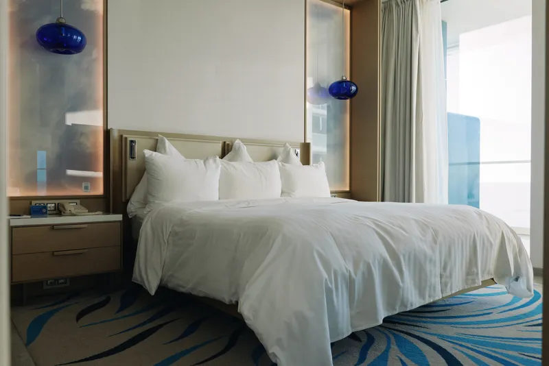 Bed, bedroom, hotel, linens, pillows, headboard, nightstand, lamps in a bright modern room with curtains, window view and soft light creating a calm, inviting space for rest and relaxation during