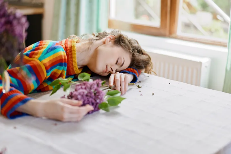 Cozy indoor scene with a woman resting at a home table, eyes closed, colorful sweater, flowers scattered, sunlit room, peaceful lifestyle moment, relaxed atmosphere