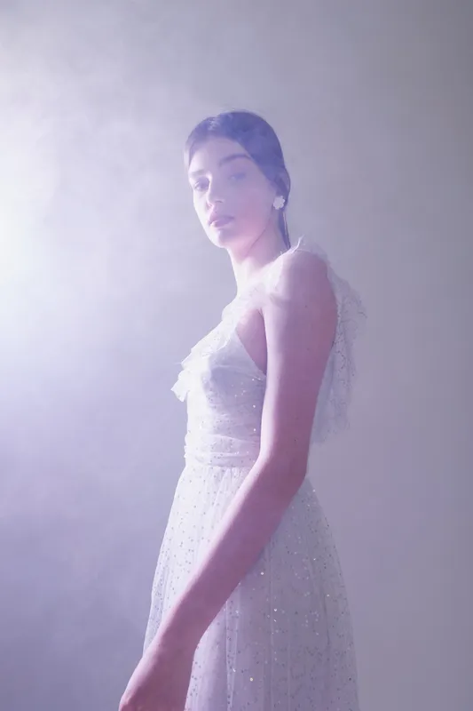 Young woman in a white elegant dress poses gracefully in soft misty light. The image highlights beauty, fashion, and delicate textures with a serene and ethereal mood.
