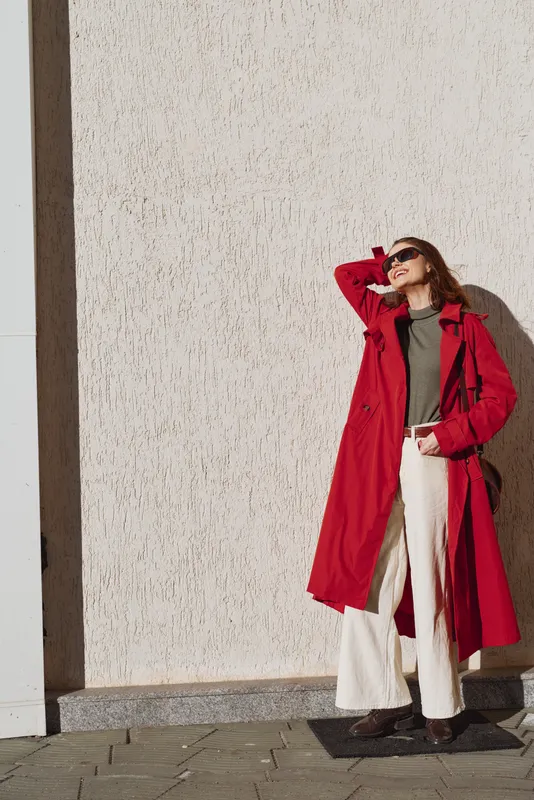 A stylish woman in a bold red coat stands against a sunlit wall, posing with one hand on her head. Outdoor fashion portrait highlighting confidence and chic street style.
