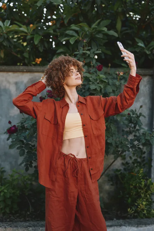 Casual woman outdoors in rust red outfit, taking a selfie, relaxed pose, sunlit greenery background, warm natural colors, candid lifestyle moment and joyful energy